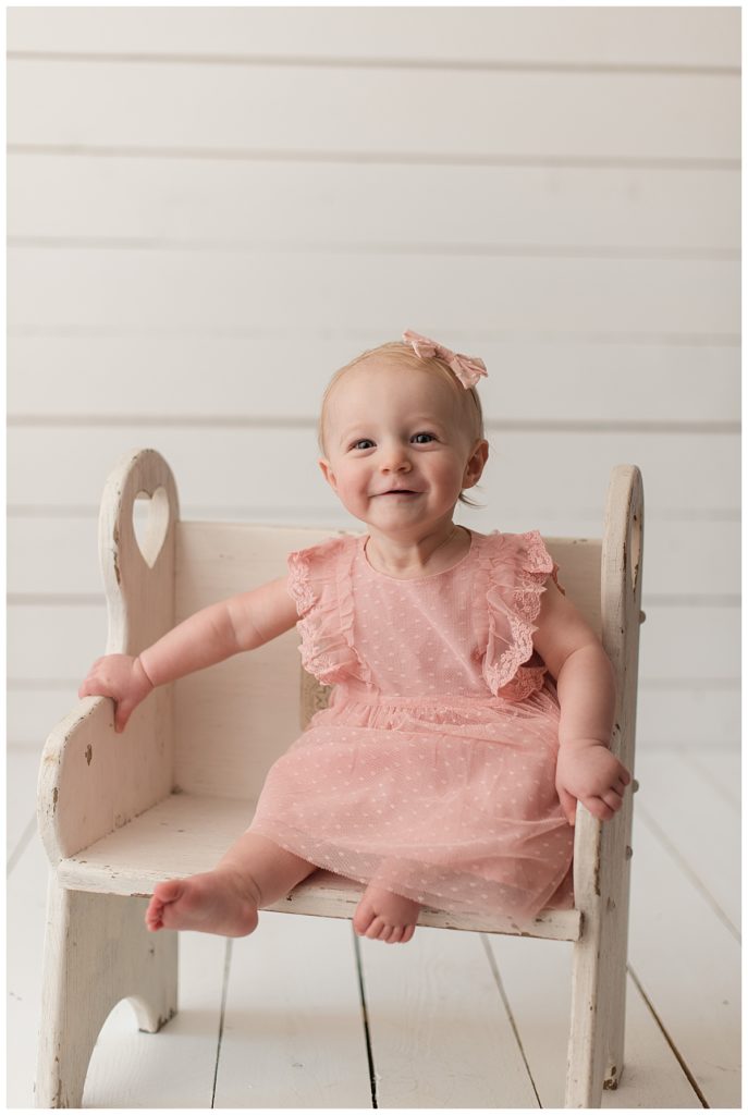 baby girl in pink dress and bow sitting on a white bench