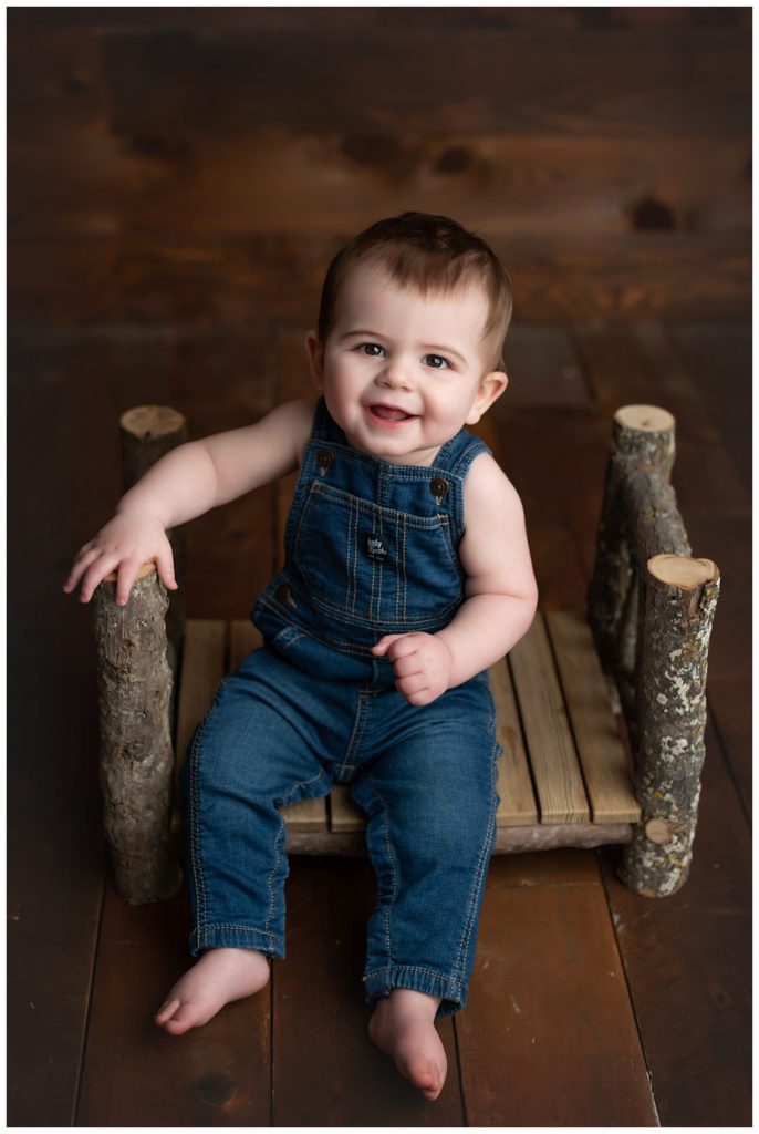 close up of baby boy in overalls on log bed on wood backdrop