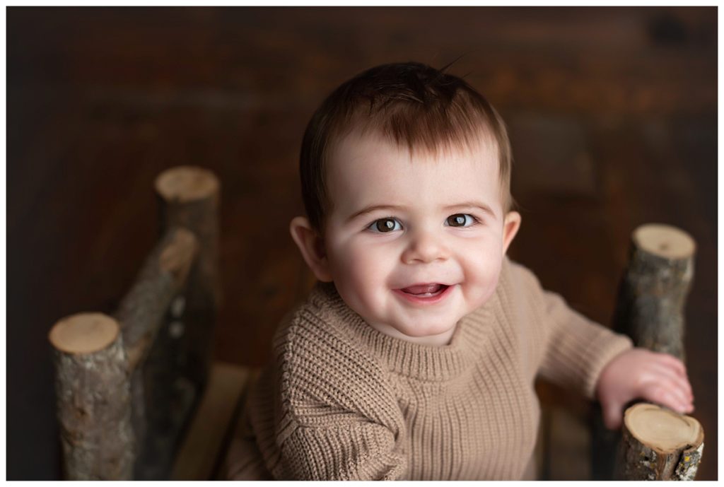 close up of baby boy in tan sweater on log bed on wood backdrop