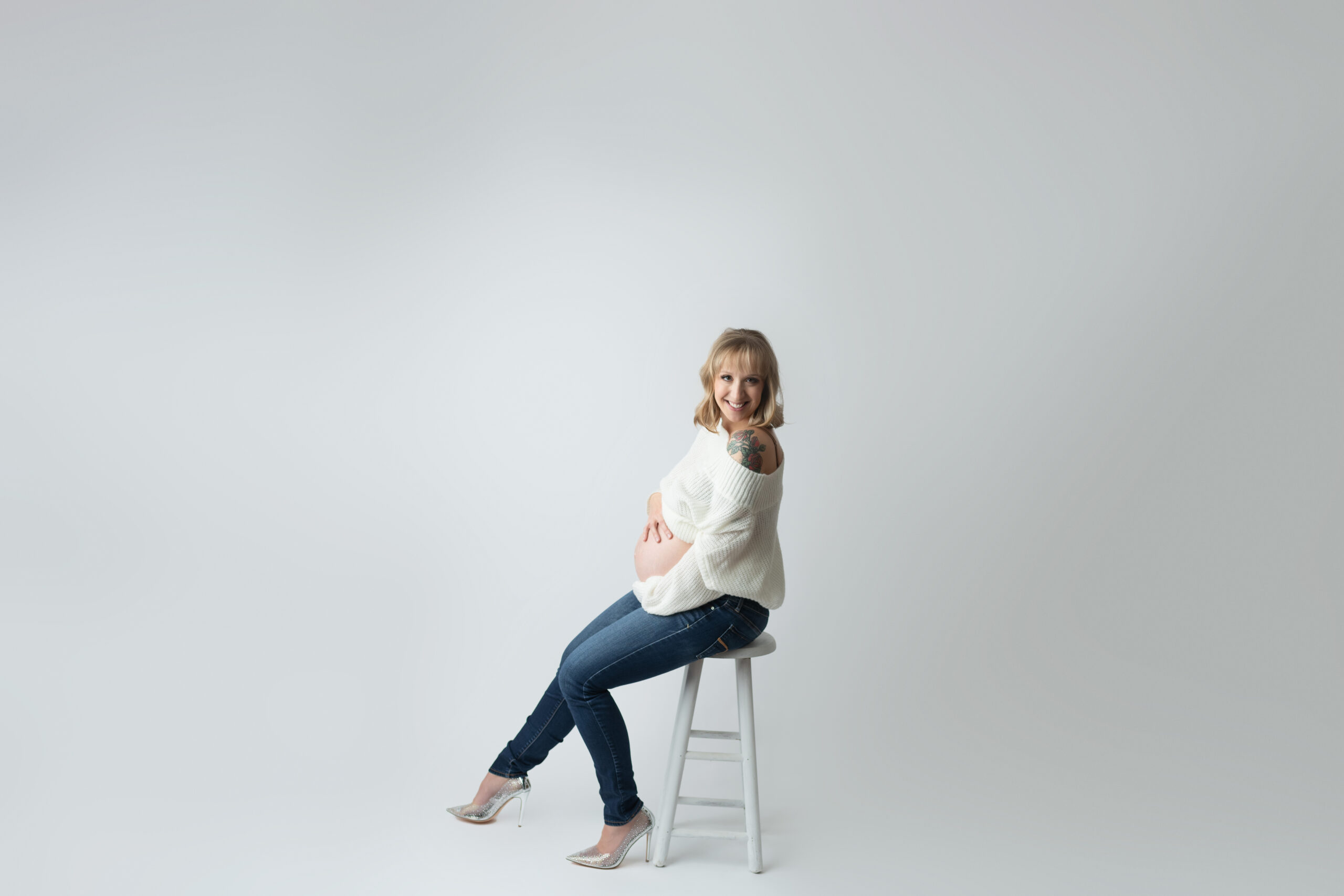 Pregnant woman showing belly wearing slouchy white sweater and jeans on white stool on white paper backdrop and sparkly heels