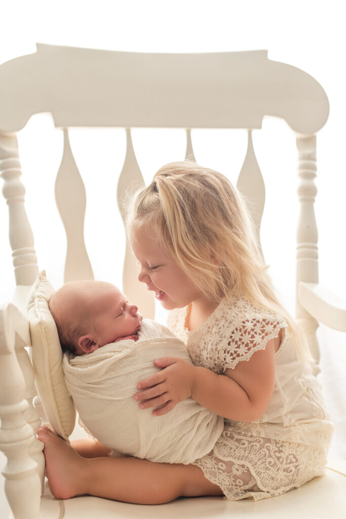 Big sister holding baby brother backlit on a white rocking chair