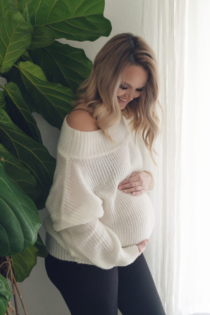 Pregnant mom in chunky white sweater in front of window and next to fiddle leaf fig