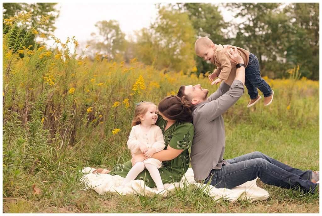 Family of four sitting on white blanket with mom looking at daughter on lap and dad tossing brother in the air and laughing