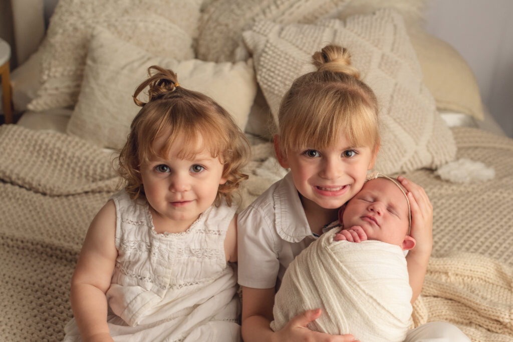 three sisters on bed together on bed while oldest sister holds the baby