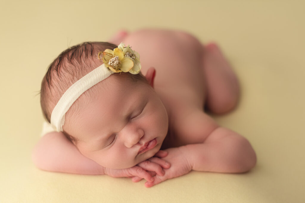 baby girl with her head on her stacked hands on yellow backdrop with yellow floral bow