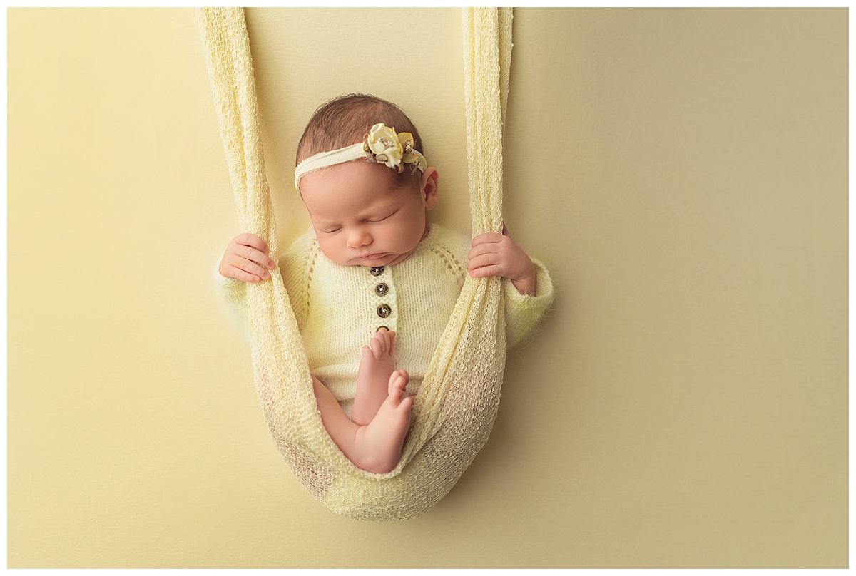 Sleeping baby girl in yellow knit long sleeve onesie with yellow tieback on yellow backdrop with yellow wrap fabric fashioned as a swing.