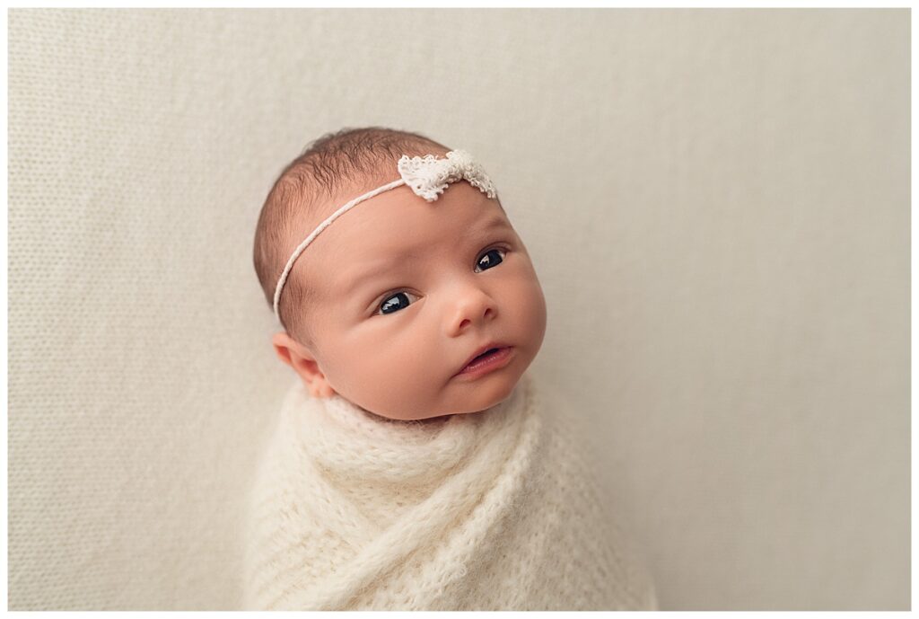 Alert newborn baby looking at camera while on white backdrop with a white bow.