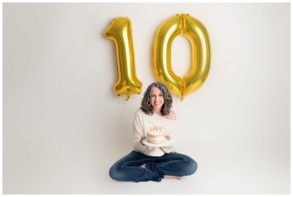 Jen sitting on the ground holding a cake with gold 10 balloons in the background